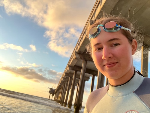 Me in goggles and a wetsuit in front of a pier on a beach