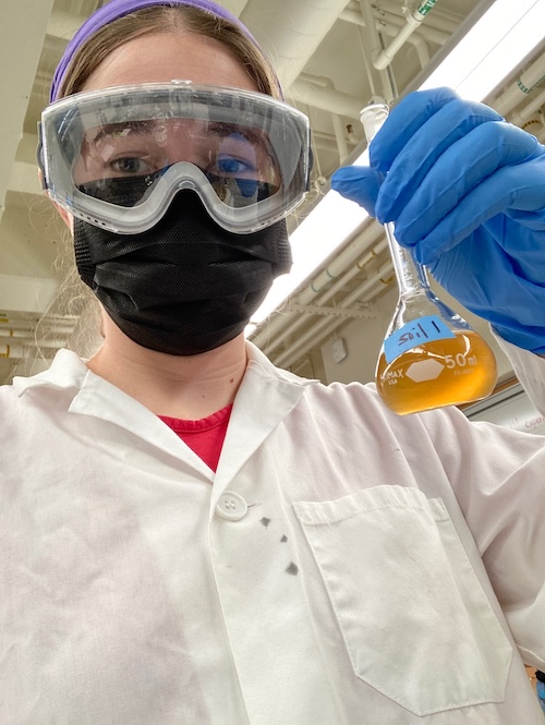 A picture of me in a lab coat, 
      goggles, and a surgical mask holding a volumetric flask of a yellow liquid